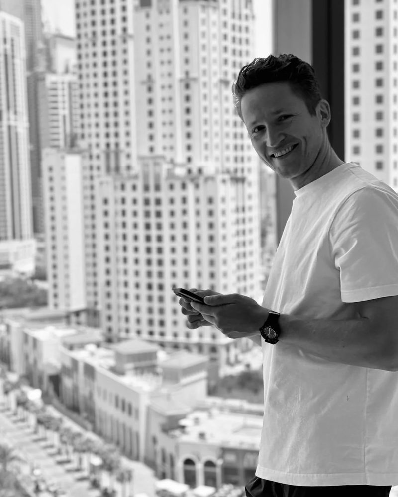 Martin Chojnicki smiling in front of a window with high-rise buildings behind him.